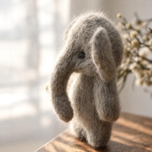 May include: A small, fuzzy, light beige needle-felted elephant figurine with a curved trunk, rounded ears, and a dark eye. The textured, soft-looking surface of the elephant shows individual fibers, creating a tactile appearance.