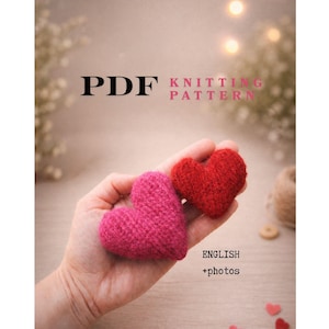 以下が含まれることがあります： PDF編み物パターン。ピンクと赤の2つの編み物ハートが手に持たれています。画像には「PDF Knitting Pattern」と「English + photos」の文字が含まれています。背景は柔らかくぼやけたボケ効果です。