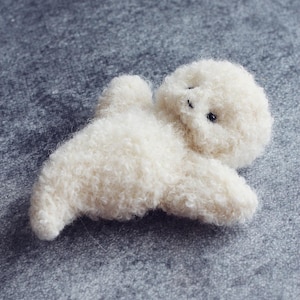 K&ouml;nnte beinhalten: Ein kleines, cremefarbenes Pl&uuml;schtier in Form eines liegenden Tieres, m&ouml;glicherweise ein Lamm oder ein Hund. Das Spielzeug hat ein flauschiges, strukturiertes Aussehen mit winzigen schwarzen Augen. Der Hintergrund ist eine einfarbige graue Oberfl&auml;che.