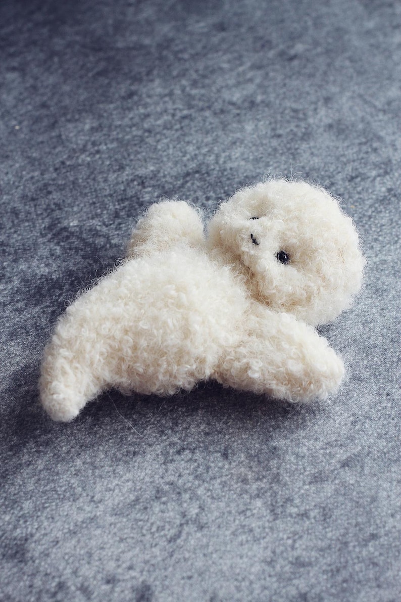 K&ouml;nnte beinhalten: Ein kleines, cremefarbenes Pl&uuml;schtier in Form eines liegenden Tieres, m&ouml;glicherweise ein Lamm oder ein Hund. Das Spielzeug hat ein flauschiges, strukturiertes Aussehen mit winzigen schwarzen Augen. Der Hintergrund ist eine einfarbige graue Oberfl&auml;che.