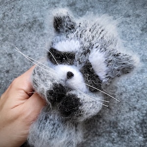 May include: A fuzzy, hand-knitted raccoon figurine crafted from grey, white, and black yarn. The raccoon features a distinct black eye mask, a small black nose, and long, thin white whiskers extending from its snout, with a soft, textured appearance.