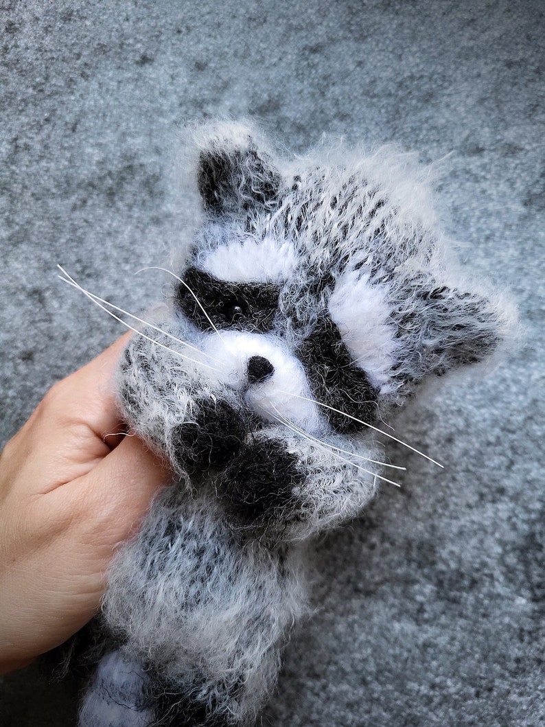 May include: A fuzzy, hand-knitted raccoon figurine crafted from grey, white, and black yarn. The raccoon features a distinct black eye mask, a small black nose, and long, thin white whiskers extending from its snout, with a soft, textured appearance.
