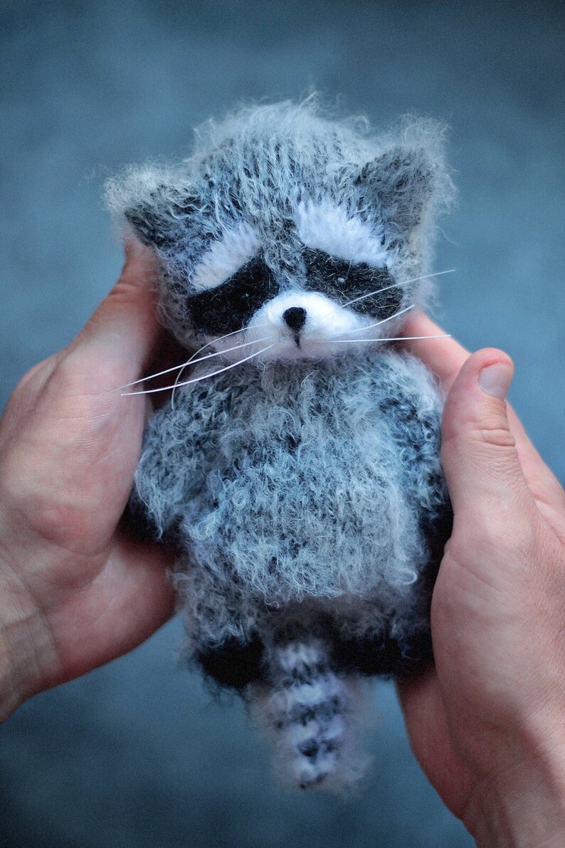 May include: A small, fuzzy, handcrafted raccoon figurine made of grey and white yarn. The raccoon features a distinct black mask around its eyes, a white muzzle, a small black nose, long white whiskers, and a striped tail with dark grey accents.