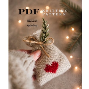 May include: A knitted bag with a red heart design, tied with twine and adorned with a sprig of greenery. The image includes the text "PDF Knitting Pattern English + photos". The bag is held against a soft, blurred background.
