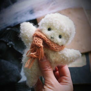 K&ouml;nnte beinhalten: Eine handgefertigte, wei&szlig;e Pl&uuml;schpuppe mit lockiger Textur, kleinen schwarzen Augen und einem aufgestickten L&auml;cheln. Die Puppe tr&auml;gt einen braunen Schal. Die Puppe wird in einer Hand gehalten.