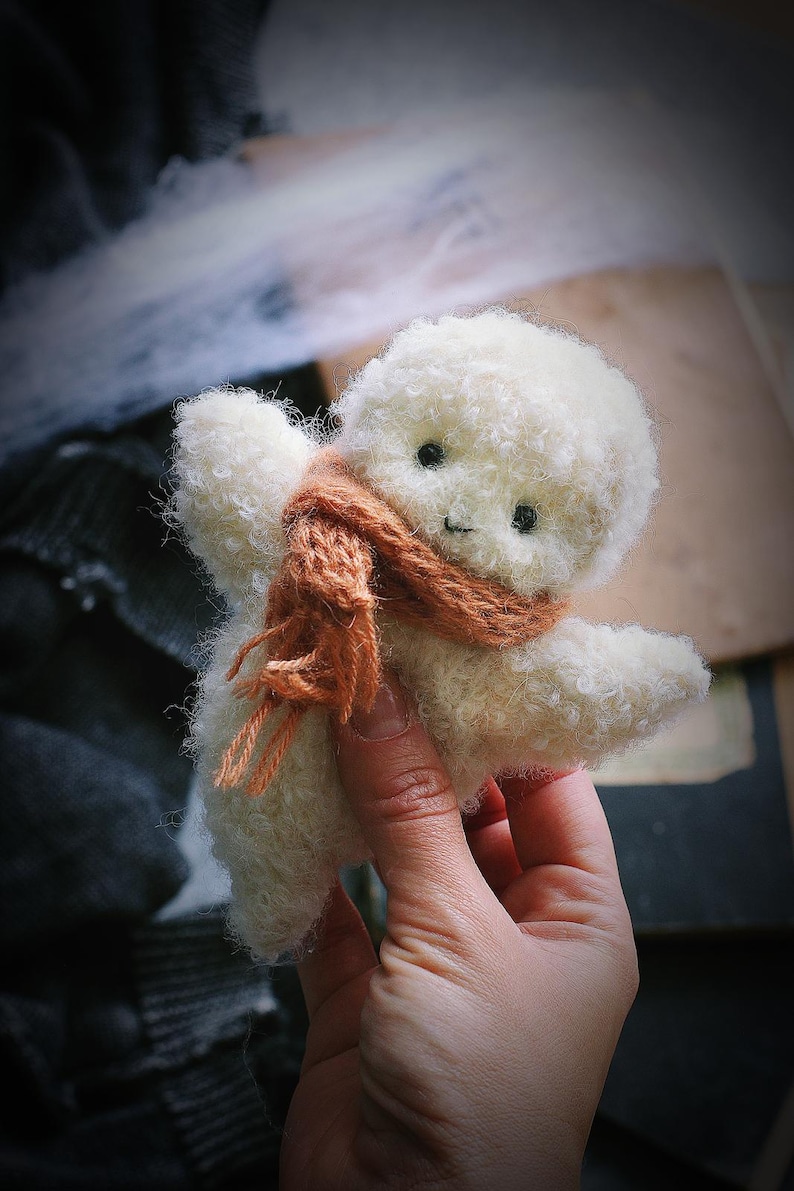 K&ouml;nnte beinhalten: Eine handgefertigte, wei&szlig;e Pl&uuml;schpuppe mit lockiger Textur, kleinen schwarzen Augen und einem aufgestickten L&auml;cheln. Die Puppe tr&auml;gt einen braunen Schal. Die Puppe wird in einer Hand gehalten.