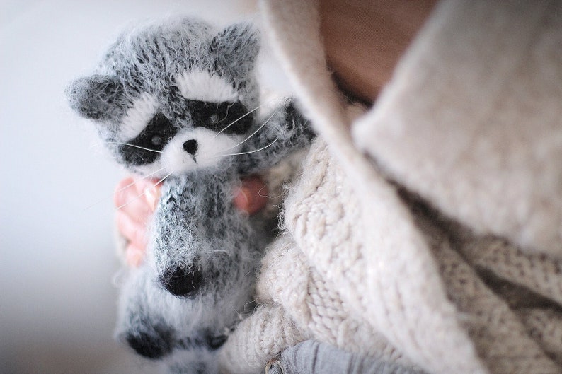 May include: A small, fuzzy, handcrafted raccoon figurine made of grey, white, and black yarn. The raccoon features a distinct black mask around its eyes, pointed ears, a small black nose, and long white whiskers extending from its snout.