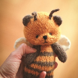 Puede incluir: Un pequeño peluche tejido a mano con forma de abeja, que presenta un cuerpo difuso a rayas naranjas y marrón oscuro, orejas de color marrón oscuro, dos antenas en la parte superior de la cabeza y dos alas color hueso en la parte posterior.
