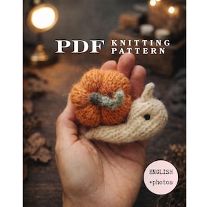Op de afbeelding: Een PDF-breipatroon met een handgebreide slak met een textuurrijke oranje pompoen als huisje en een crèmekleurig lichaam. De tekst op de afbeelding luidt PDF KNITTING PATTERN en ENGLISH +photos. De slak heeft een klein donker oog en een groen steeltje.