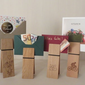 May include: Five wooden card holders with different engraved designs. Each holder has a black elastic band and is holding a card or envelope. The cards include a wedding card, a gift certificate, a card that says "Alles Gute!" and an envelope with cash.
