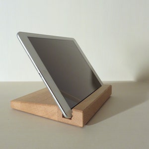 Pode incluir: Um suporte de madeira com um tablet apoiado em ângulo. O suporte é de madeira marrom claro e o tablet é prateado com uma tela preta.