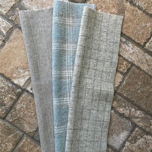 May include: Three folded fabric swatches in varying patterns. The first is a light gray with a small, textured pattern. The second is a light blue plaid. The third is a gray plaid. The fabrics are displayed on a brown stone surface.