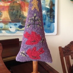 Puede incluir: Un árbol decorativo en forma de cono, morado, hecho a mano, con base de madera. El árbol presenta detalles bordados, incluyendo una estrella dorada, un árbol verde y dos diseños florales rojos. El árbol mide aproximadamente 25 cm de alto.