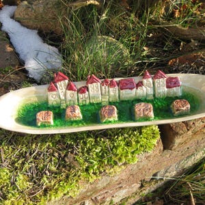 Könnte beinhalten: Eine dekorative Keramikschale mit einer Miniatur-Dorfszene. Das Dorf hat weiße Gebäude mit roten Dächern, die auf einer grünen Basis stehen. Die Schale enthält auch braune, quadratische Elemente. Die Szene spielt im Freien.