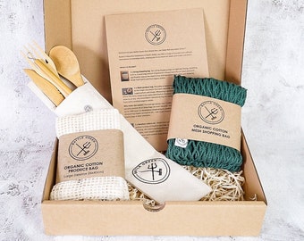 May include: A brown cardboard box with a green organic cotton mesh shopping bag, a white organic cotton produce bag, and a set of bamboo cutlery. The box is filled with brown packing material. The text on the bags reads "Organic Cotton Mesh Shopping Bag" and "Organic Cotton Produce Bag".