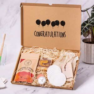 Puede incluir: Una caja de cartón marrón con las palabras "CONGRATULATIONS" impresas en la tapa. La caja está abierta y contiene un set de regalo con una mascarilla facial, jabón, bálsamo labial, discos de algodón y una bolsa de malla reutilizable. El set de regalo es perfecto para una ocasión especial.