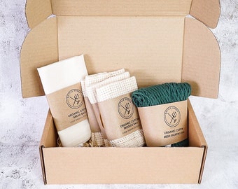 May include: A brown cardboard box with a lid open, revealing a set of reusable shopping bags. The set includes a large white cotton bread bag, two green mesh produce bags, and a green cotton string bag. The bags are labeled with the text "Battle Green" and "Organic Cotton".