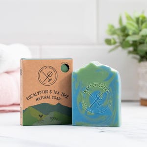 Op de afbeelding: Een natuurlijke zeep met blauwe en groene wervelingen, naast de kartonnen verpakking. De doos toont "Eucalyptus & Tea Tree Natural Soap" en een logo. De zeep weegt 100g.