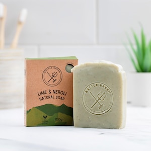 May include: A green bar of natural soap and its box, both labelled "Lime & Neroli" with the "Battle Green" logo. The box is brown with green accents and the soap is 100g. A small plant and toothbrushes are in the background.