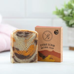 May include: A bar of citrus scrub natural soap with a brown and orange swirl pattern, next to a brown box. The soap has a circular logo with the words "Battle Green" and a trident. The box reads "Citrus Scrub Natural Soap" and "100g".