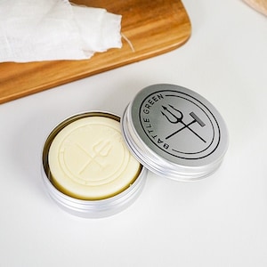 Könnte beinhalten: Geöffnete, kleine, silberne Dose mit einem runden, hellgelben Seifenstück. Die Seife hat ein Dreizack-Design. Der Deckel, neben der Dose, trägt die Aufschrift "BATTLE GREEN" und ein Dreizack- und Kamm-Design.