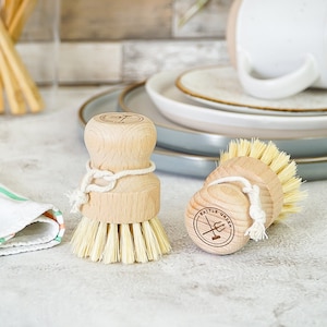 Könnte beinhalten: Zwei Holz-Spülbürsten mit Naturborsten. Die Bürsten haben eine weiße Schnur, die um die Mitte gebunden ist, und einen Holzgriff mit den Worten "Battle Green" und einem Logo, das auf der Unterseite aufgedruckt ist.