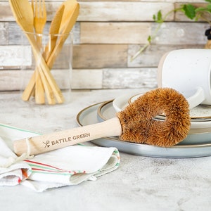 Peut inclure: Une brosse à vaisselle en fibre de noix de coco brune avec un manche en bois. Le manche est gravé du texte "BATTLE GREEN" et d'un logo de couverts croisés. La brosse est posée sur une serviette rayée blanche et verte à côté d'une tasse en céramique blanche et d'un ensemble d'ustensiles en bois.