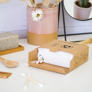 May include: A box of 100 biodegradable cotton swabs with bamboo sticks. The box is made of recycled cardboard and has a green recycling symbol on it. The box is open and the cotton swabs are visible inside.
