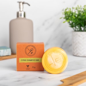以下が含まれることがあります： 丸い黄色のシトラスシャンプーバーと、"Battle Green Citrus Shampoo Bar"の文字とトライデントのロゴが入ったオレンジ色の箱。箱には「55g」とも記載されています。灰色の石鹸ディスペンサーと小さな植物が背景にあります。