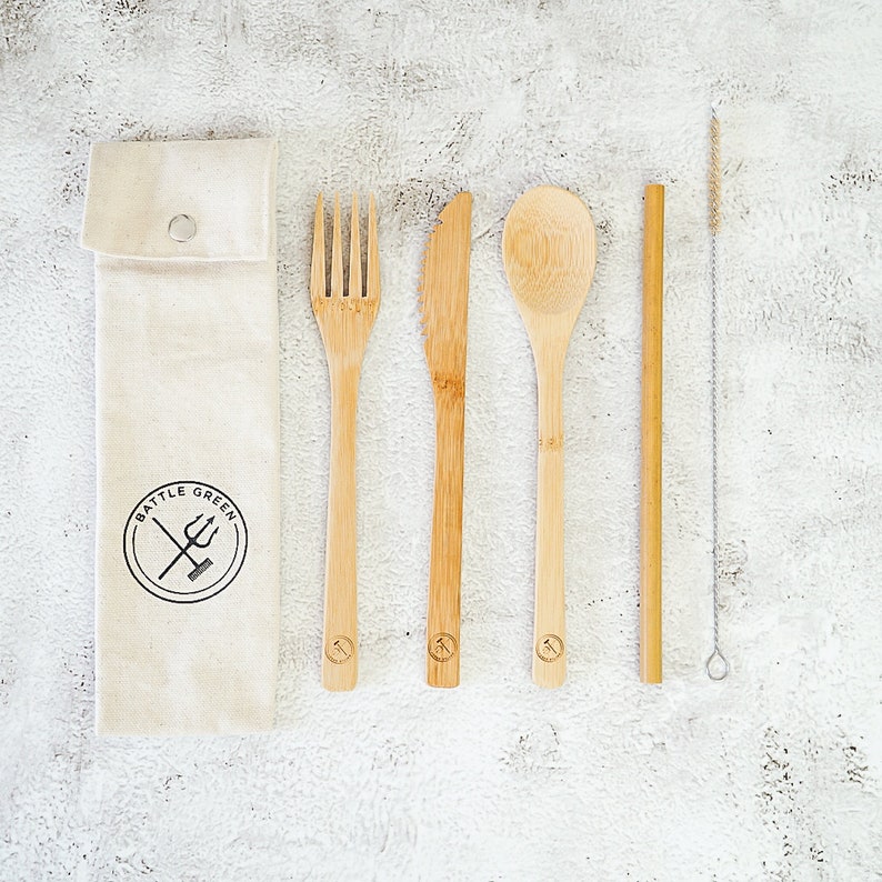 Puede incluir: Un juego de cinco utensilios de bamb&uacute;, que incluye un tenedor, un cuchillo, una cuchara, una pajita y un cepillo de limpieza, est&aacute;n dispuestos sobre un fondo blanco. Los utensilios est&aacute;n empaquetados en una bolsa de lona blanca con el logotipo "Battle Green" impreso en ella.
