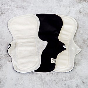 May include: Three reusable cloth menstrual pads, one black and two white. The pads are folded and stacked on top of each other. The black pad has a small white label with text on it.