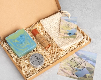 May include: A cardboard gift box containing a blue and green marbled soap bar, a brown cylindrical lip balm, a beige woven exfoliating cloth, and a small round metal tin. Each item features a logo with a trident and the text Battle Green.