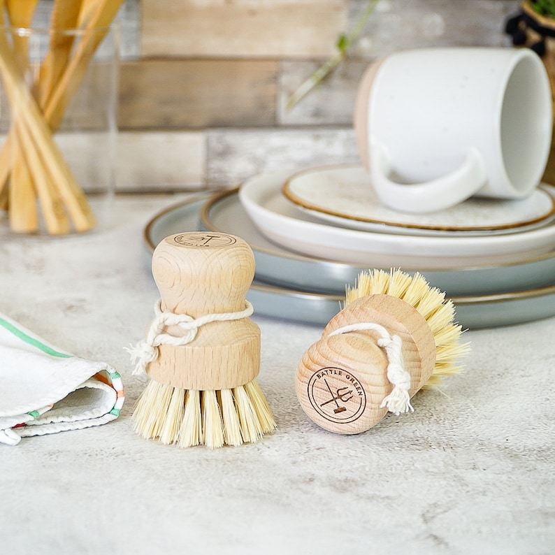May include: Two wooden dish brushes with natural bristles. The brushes have a white string tied around the middle and a brown logo that says "Battle Green" with a crossed trident and broom design.