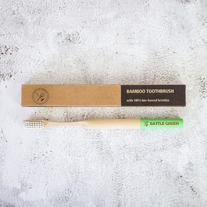 Puede incluir: Un cepillo de dientes de bamb&uacute; con cerdas blancas y una etiqueta verde que dice "BATTLE GREEN". El cepillo de dientes est&aacute; empaquetado en una caja de cart&oacute;n marr&oacute;n con el texto "BAMBOO TOOTHBRUSH with 100% bio-based bristles".