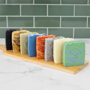 May include: A row of eight rectangular soap bars in various colours including black, marbled tan, sage green, blue, orange, grey, cream, and a green and blue swirl. Each bar features a circular stamp with the text Battle Green and a trident icon.