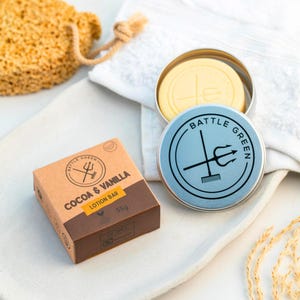 Op de afbeelding: Een bruine en bruin-gele cacao- en vanille-lotionbar in een kartonnen doos, naast een lichtblauwe blik met de woorden "Battle Green" en een drietandlogo. Een gele lotionbar zit in een zilveren blik. Een natuurlijke spons en witte handdoek staan op de achtergrond.