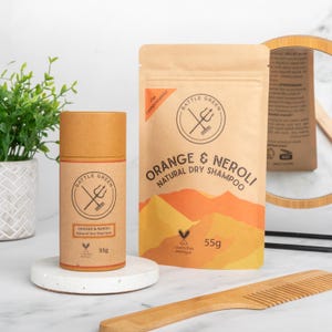May include: A brown cardboard tube and pouch of "Orange & Neroli Natural Dry Shampoo" by Battle Green. The pouch features an orange and yellow mountain design. The tube and pouch are on a white surface with a comb and mirror in the background. The product weighs 55g.