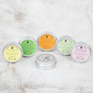 Op de afbeelding: Een verzameling van zes kleine, ronde lipbalsemblikjes met kleurrijke etiketten. De etiketten tonen smaken zoals Vanille, Limoen, Mandarijn, Pepermunt en Roos. Een zevende blikje met een logo in het midden.