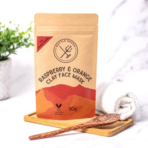 May include: A brown paper pouch with a red and orange design. The pouch contains a raspberry and orange clay face mask. The pouch is labelled "Battle Green" and "I'm compostable!" The pouch also says "Raspberry & Orange Clay Face Mask" and "50g".