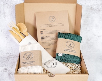 May include: A brown cardboard box with a green and white organic cotton mesh shopping bag, a white organic cotton produce bag, and a set of wooden utensils. The box is filled with brown packing material. The text on the bags reads "Organic Cotton Mesh Shopping Bag" and "Organic Cotton Produce Bag".
