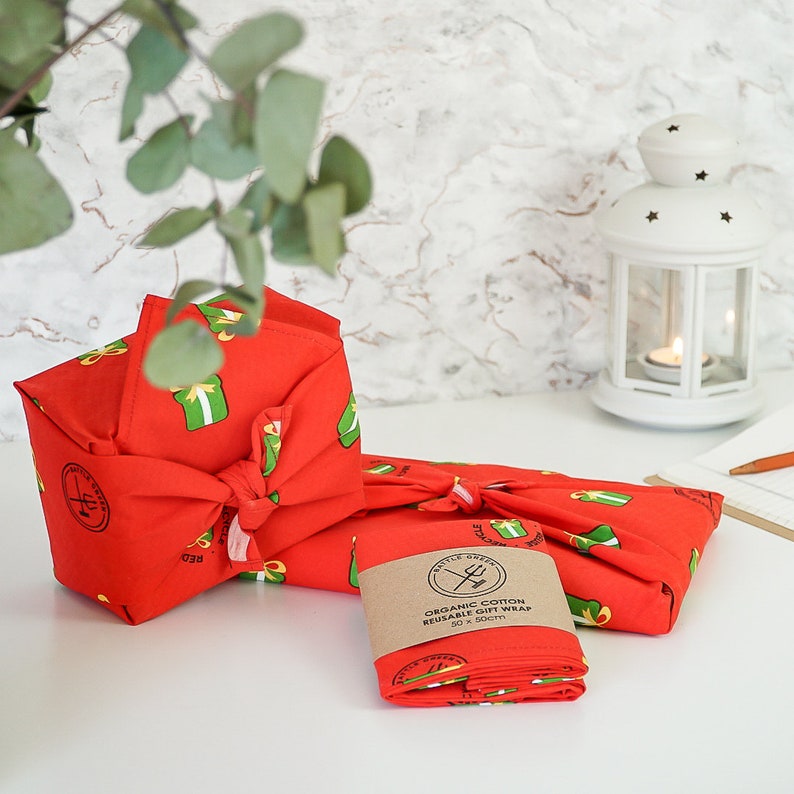Puede incluir: Dos telas de envolver regalos reutilizables rojas con un patr&oacute;n de regalos verdes. La tela est&aacute; hecha de algod&oacute;n org&aacute;nico y mide 50 x 50 cm. El texto "Organic Cotton Reusable Gift Wrap 50 x 50cm" est&aacute; impreso en una etiqueta marr&oacute;n.