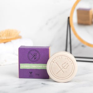 May include: A round, pale conditioner bar and a purple box, both labelled "Battle Green." The box states "Lavender Conditioner Bar" and "55g." The background features a mirror and a brush.