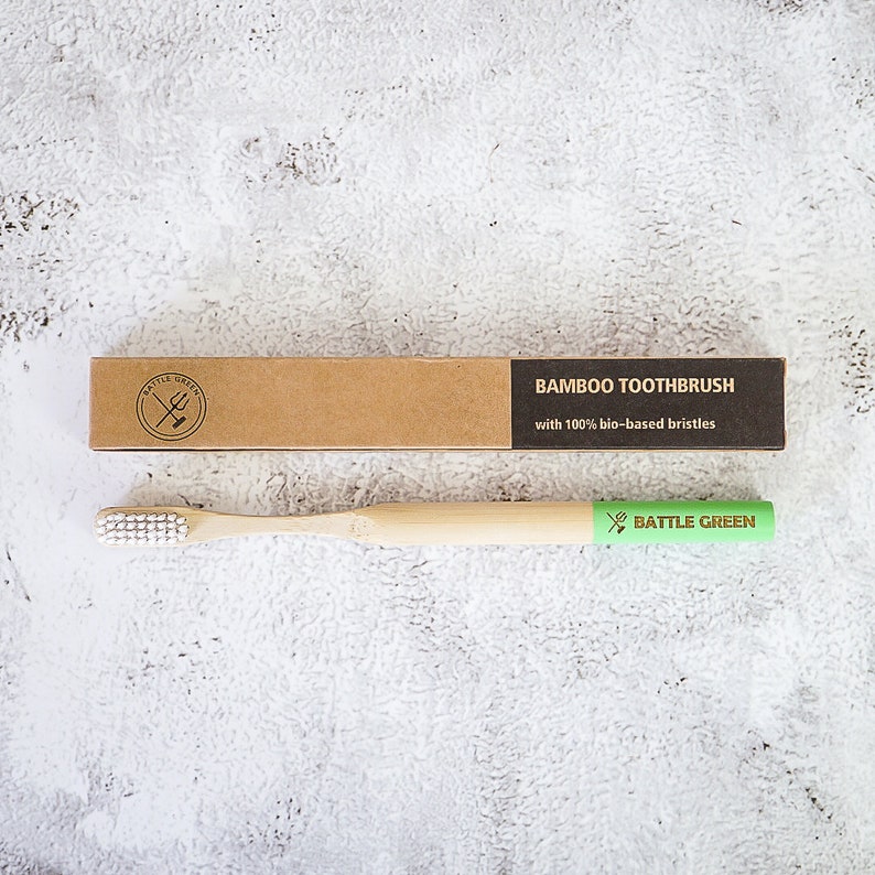 Puede incluir: Un cepillo de dientes de bamb&uacute; con cerdas blancas y una etiqueta verde que dice "BATTLE GREEN". El cepillo de dientes est&aacute; empaquetado en una caja de cart&oacute;n marr&oacute;n con el texto "BAMBOO TOOTHBRUSH with 100% bio-based bristles".