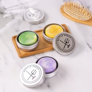 Puede incluir: Cuatro latas plateadas con tapas que contienen barras s&oacute;lidas de jab&oacute;n de diferentes colores. Las tapas tienen un logotipo en blanco y negro con el texto "BATTLE GREEN" y un tridente y un cepillo.