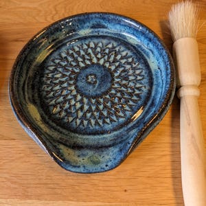 May include: A handmade blue ceramic grater with a floral pattern in the centre. The grater has a curved edge and is accompanied by a wooden-handled brush with white bristles. The grater is approximately 20 cm in diameter.