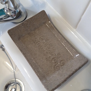 Op de afbeelding: Een lichtbruine keramische zeepbak met een textuur. De bak is rechthoekig met afgeronde hoeken en heeft een decoratief ontwerp.