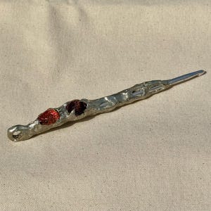 以下が含まれることがあります： ループが付いた銀色の杖型のオブジェクト。杖は片方の端にループがあり、先端が尖っています。杖はテクスチャ加工された表面を持ち、2つの赤い装飾が施されています。ベージュ色の布の表面に表示されています。