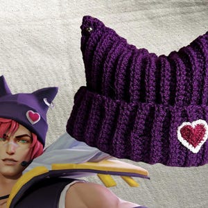May include: A deep purple crochet beanie hat with cat ears and a heart-shaped patch. The hat has a ribbed texture and a silver hoop earring. A similar hat is worn by a character in the image.