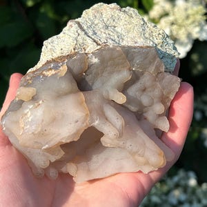 May include: A natural mineral specimen with a layered appearance. The top section is a rough, textured, light green colour, while the bottom is a translucent, off-white colour with a bubbly texture. Held in a hand.