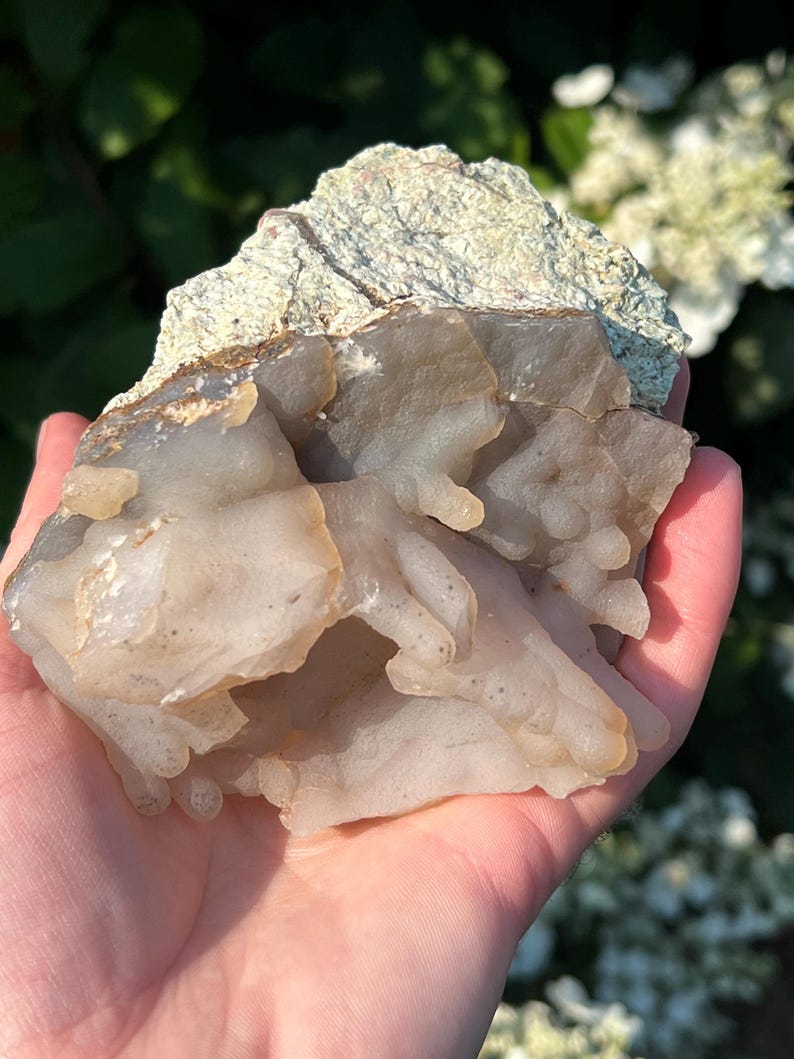 May include: A natural mineral specimen with a layered appearance. The top section is a rough, textured, light green colour, while the bottom is a translucent, off-white colour with a bubbly texture. Held in a hand.